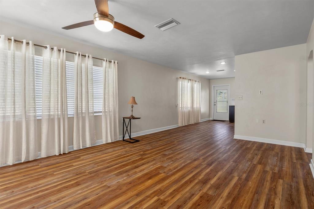 A spacious living room features wood-look flooring, a ceiling fan, and large windows with curtains — 1213 8TH STREET NW, WINTER HAVEN, FL