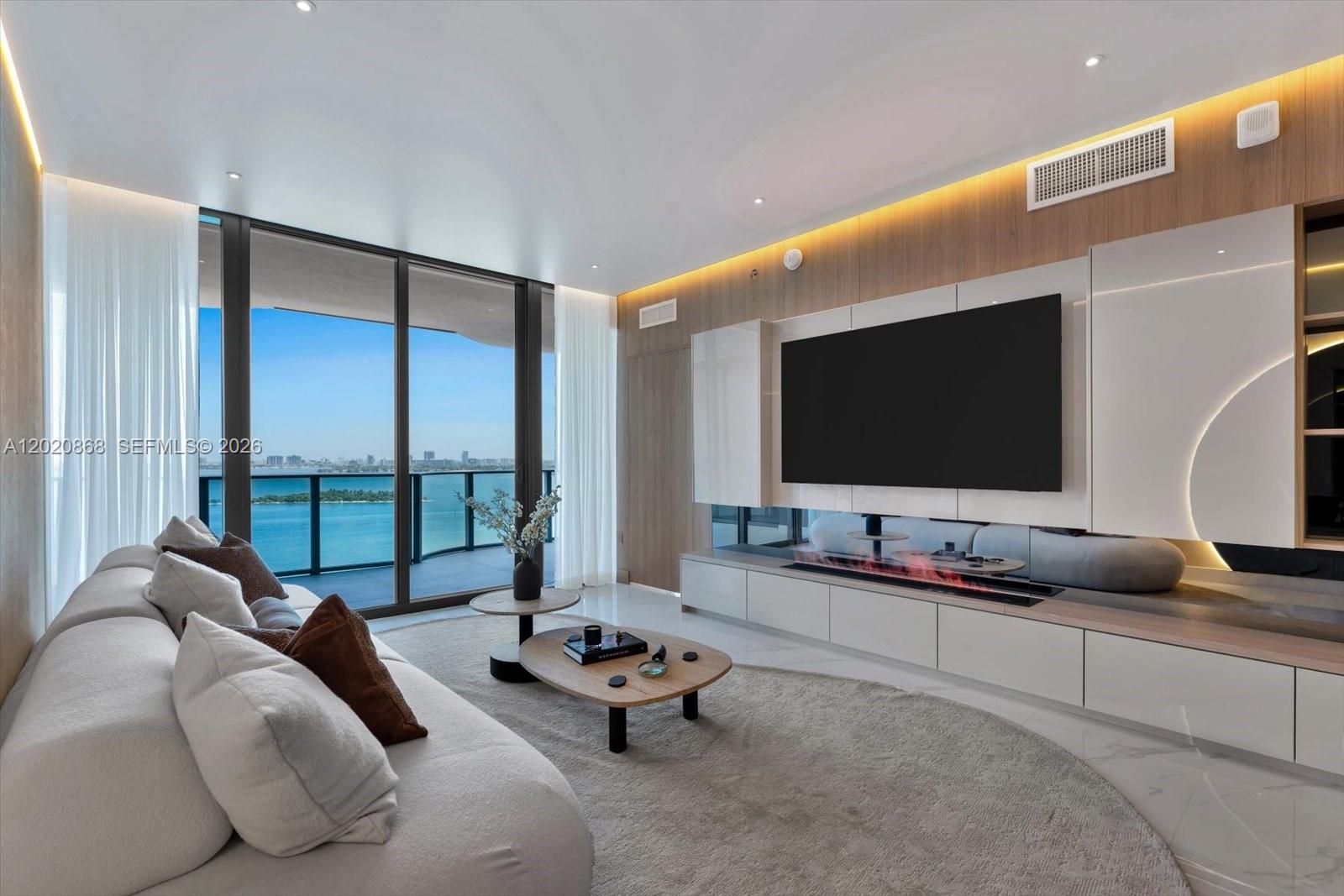 A modern living room features a large television, linear fireplace, and floor-to-ceiling windows overlooking water — 700 NE 24th St # 1602, Miami FL 33137, MIAMI, FL