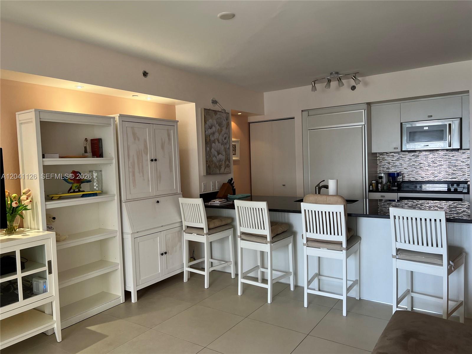 A kitchen features white cabinetry, a dark countertop, and four white bar stools — HALLANDALE BEACH, FL