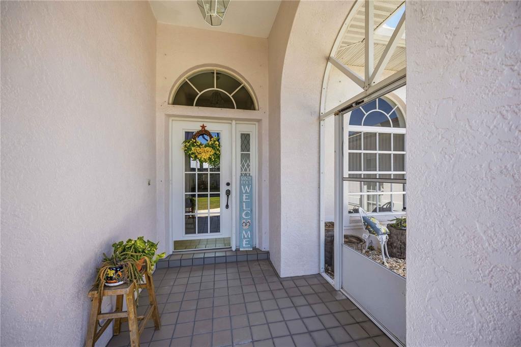 A covered entryway features a white front door with a sidelight, tile flooring, and plants — 23530 CLAY AVENUE, PORT CHARLOTTE, FL