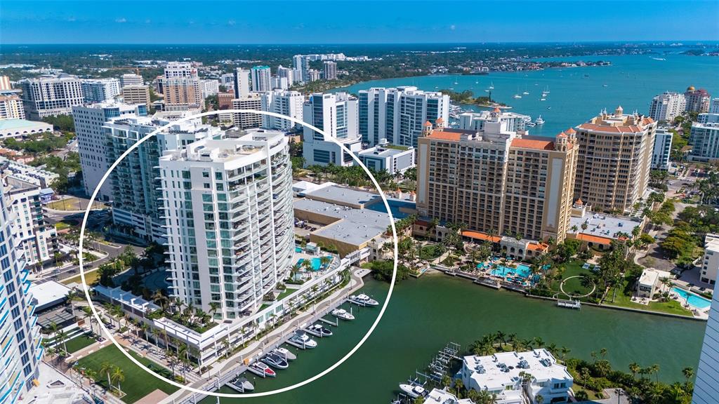 An aerial view shows a modern white high-rise residential building situated along a waterfront marina — 401 QUAY COMMONS UNIT 505, SARASOTA, FL
