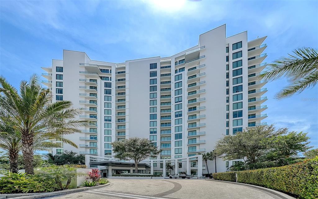 A multi-story white residential building features balconies and a paved circular driveway with landscaping — 401 QUAY COMMONS UNIT 505, SARASOTA, FL