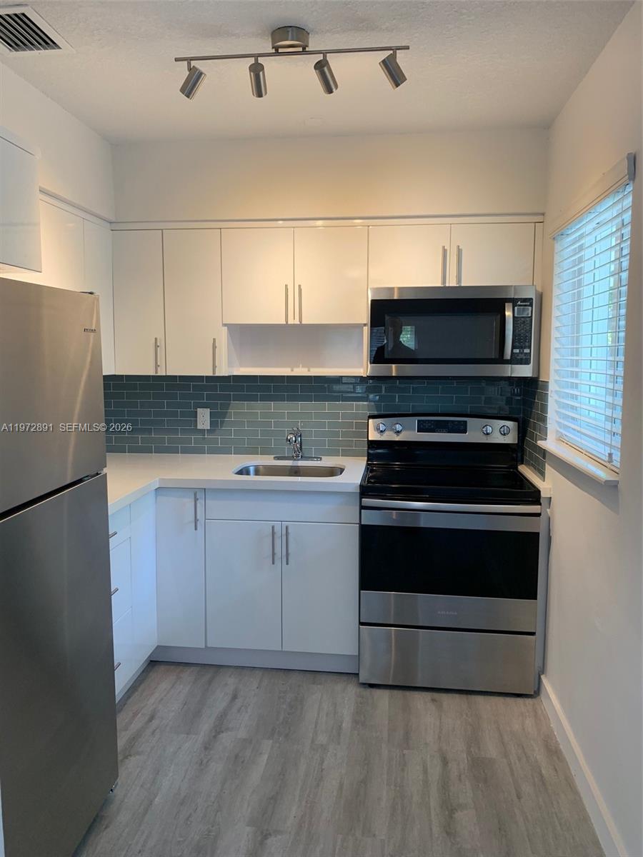 A kitchen features white cabinets, stainless steel appliances, a teal subway tile backsplash, and wood-look flooring — FORT LAUDERDALE, FL