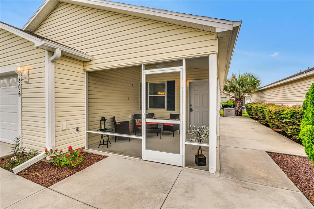 A light-colored house exterior features a screened-in porch with patio furniture and a concrete walkway — 1806 QUEENSBURY TERRACE, THE VILLAGES, FL