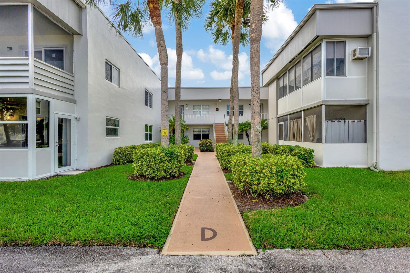 A concrete walkway leads between two multi-story apartment buildings with palm trees and landscaping — 185 Normandy # D, Delray Beach FL 33484, DELRAY BEACH, FL