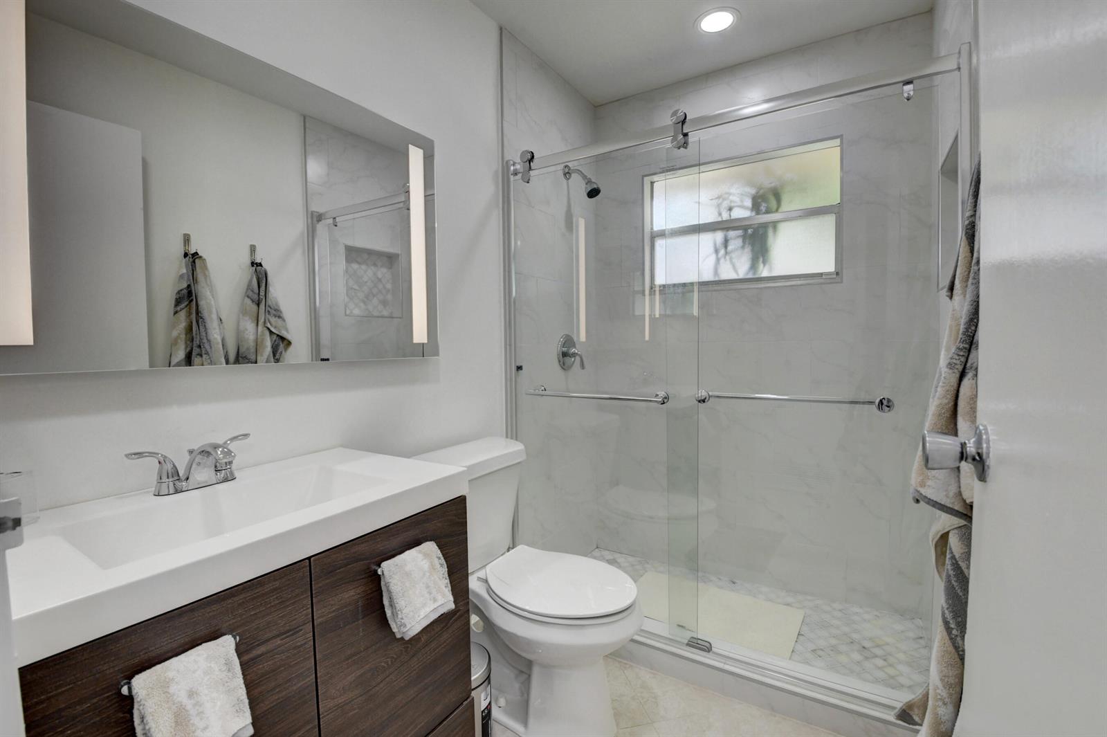 A modern bathroom features a wood vanity, white toilet, and a glass-enclosed walk-in shower — 468 Saxony Way, Delray Beach FL 33446, DELRAY BEACH, FL