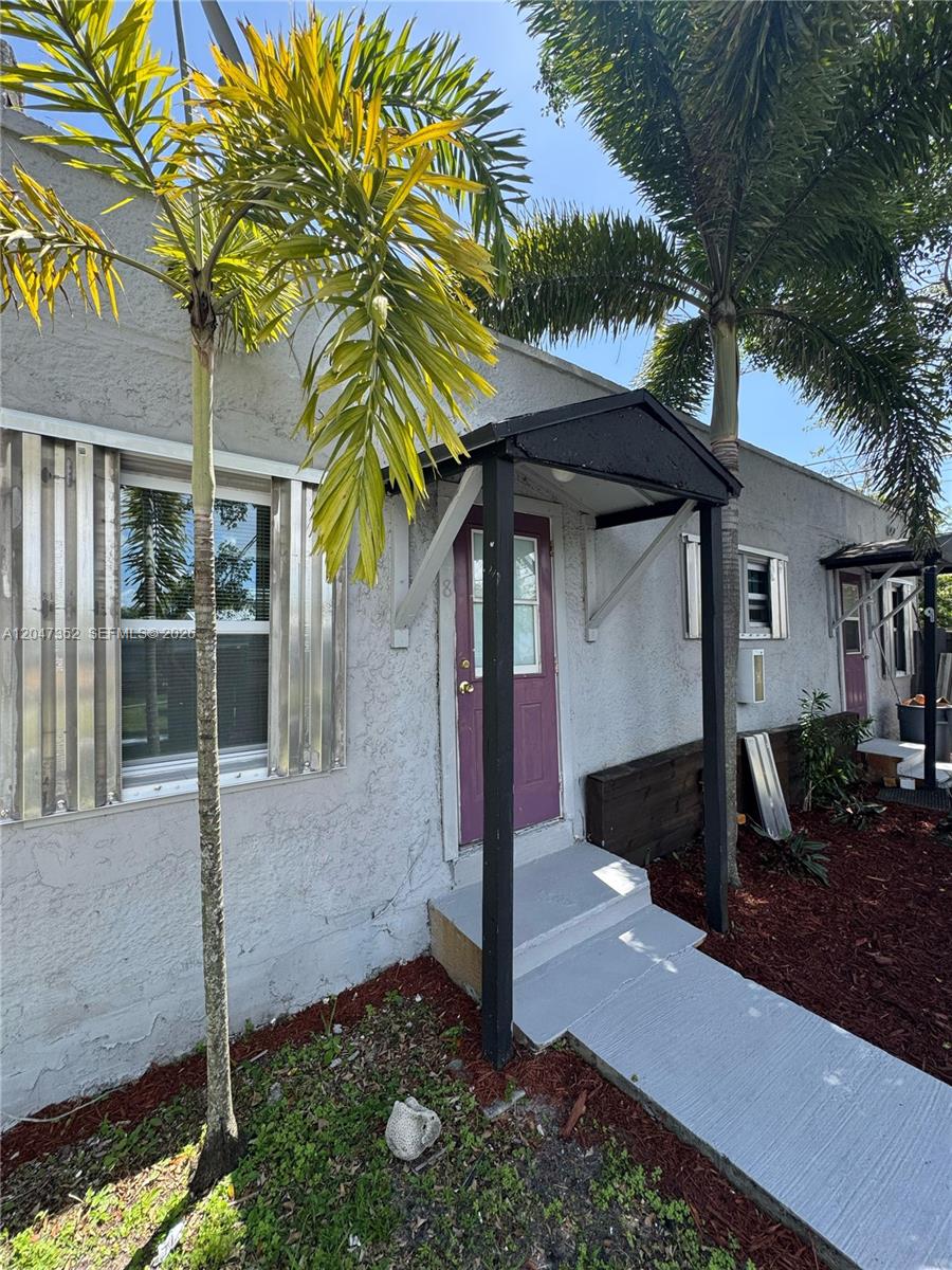 The exterior of a gray stucco building features a purple door and palm trees — DANIA BEACH, FL