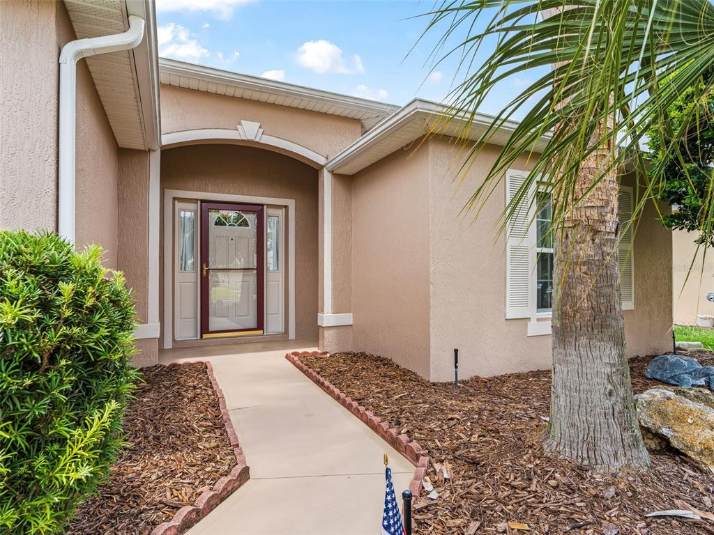 A tan stucco house exterior features a covered entryway, a red front door, and landscaping — 15360 SW 15TH TERRACE ROAD, OCALA, FL