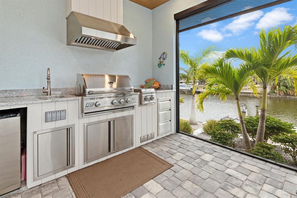 An outdoor kitchen with stainless steel appliances and granite countertops overlooks a canal and palm trees — 15650 MEACHAM CIRCLE, PORT CHARLOTTE, FL