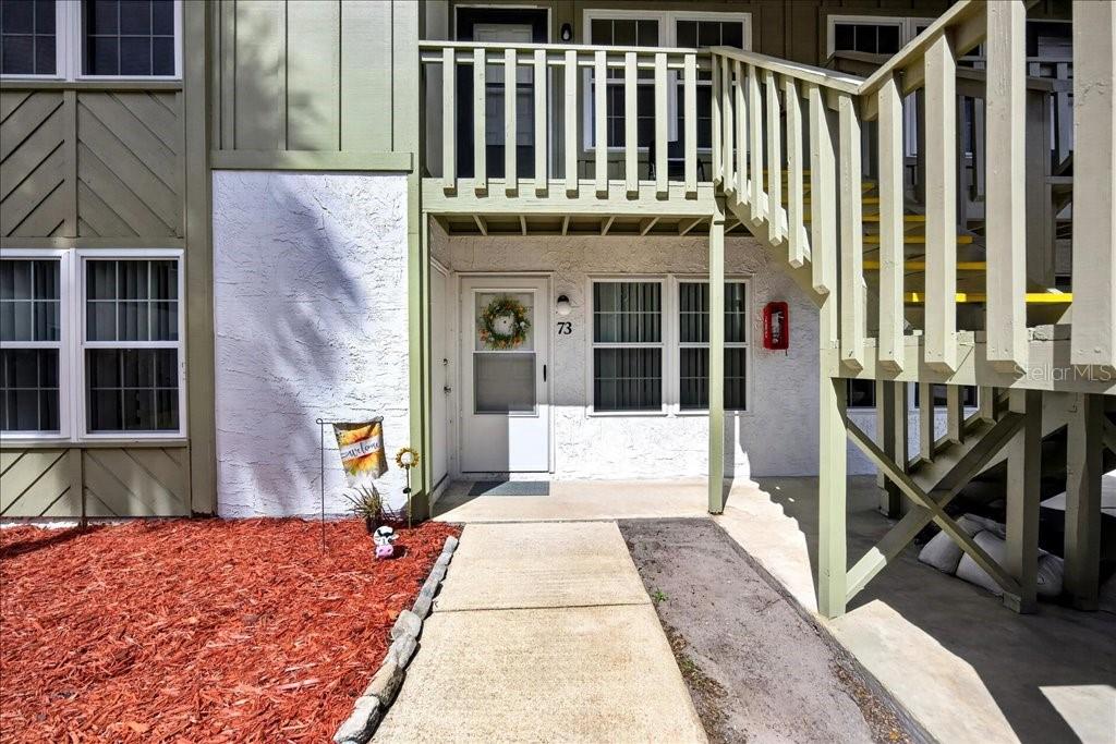 A ground-level apartment entrance with a concrete walkway, white stucco walls, and wooden stairs — 840 CENTER AVENUE UNIT 730, DAYTONA BEACH, FL