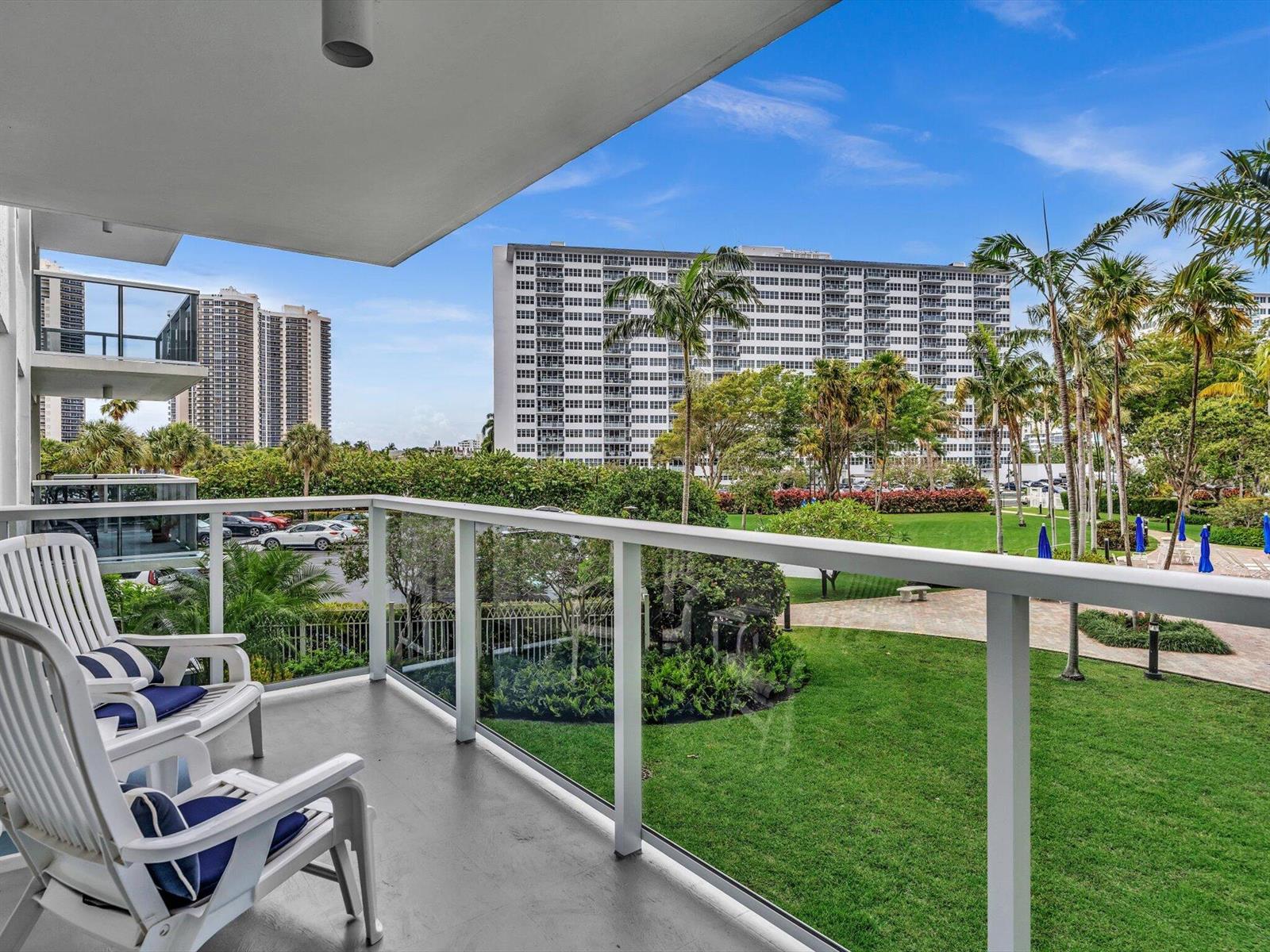 A balcony with white chairs overlooks a grassy courtyard, palm trees, and high-rise apartment buildings — 3300 NE 36th Street # 217, FL 33308, FL