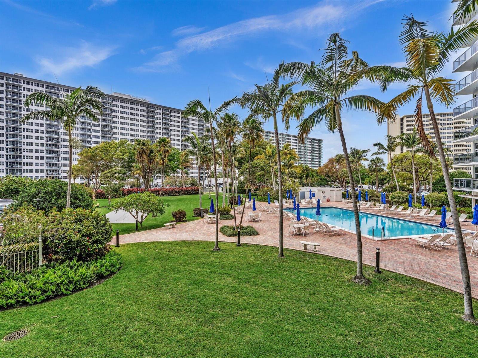A swimming pool with blue umbrellas and lounge chairs sits beside a large apartment building — 3300 NE 36th Street # 217, FL 33308, FL