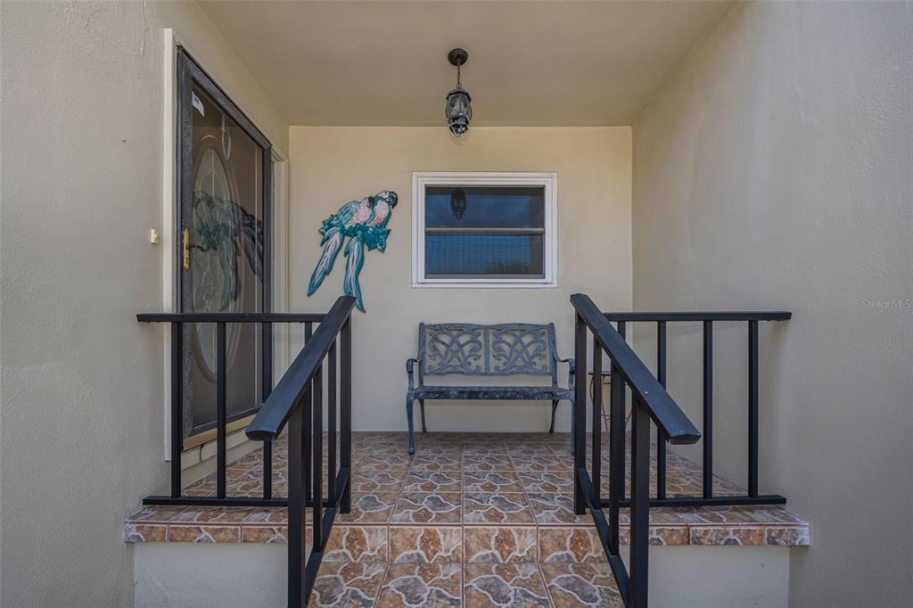 A covered entryway features tiled steps, black railings, a metal bench, and a parrot wall decoration — 2700 BAYSHORE BOULEVARD UNIT 525, DUNEDIN, FL