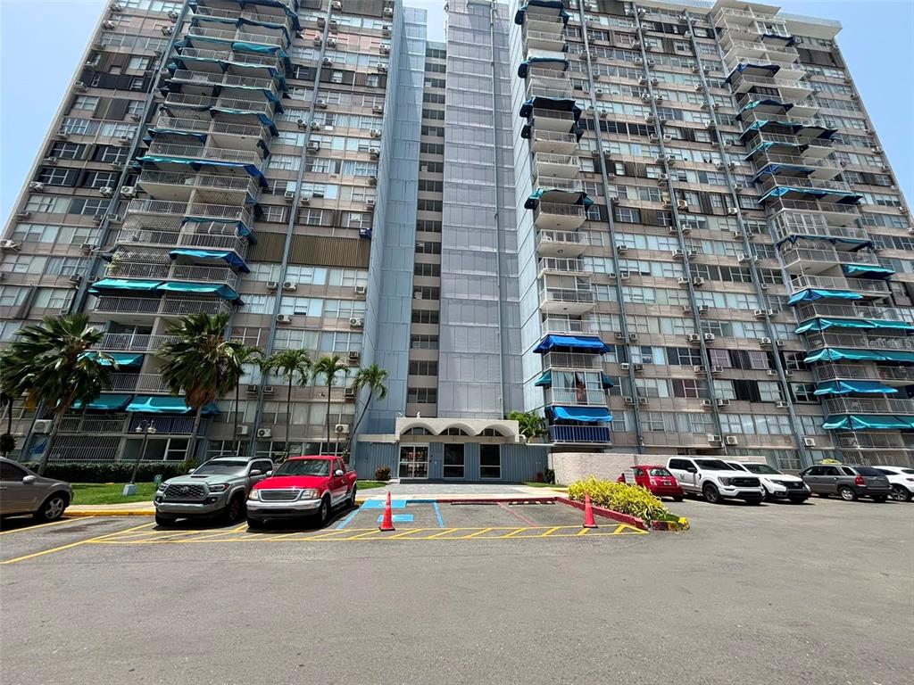 A tall residential apartment building with blue balcony awnings stands behind a parking lot — 1005 Apt CONDOMINIO MUNDO FELIZ (10MO PISO - BALDORIOTY TOW UNIT 1005, CAROLINA, PR