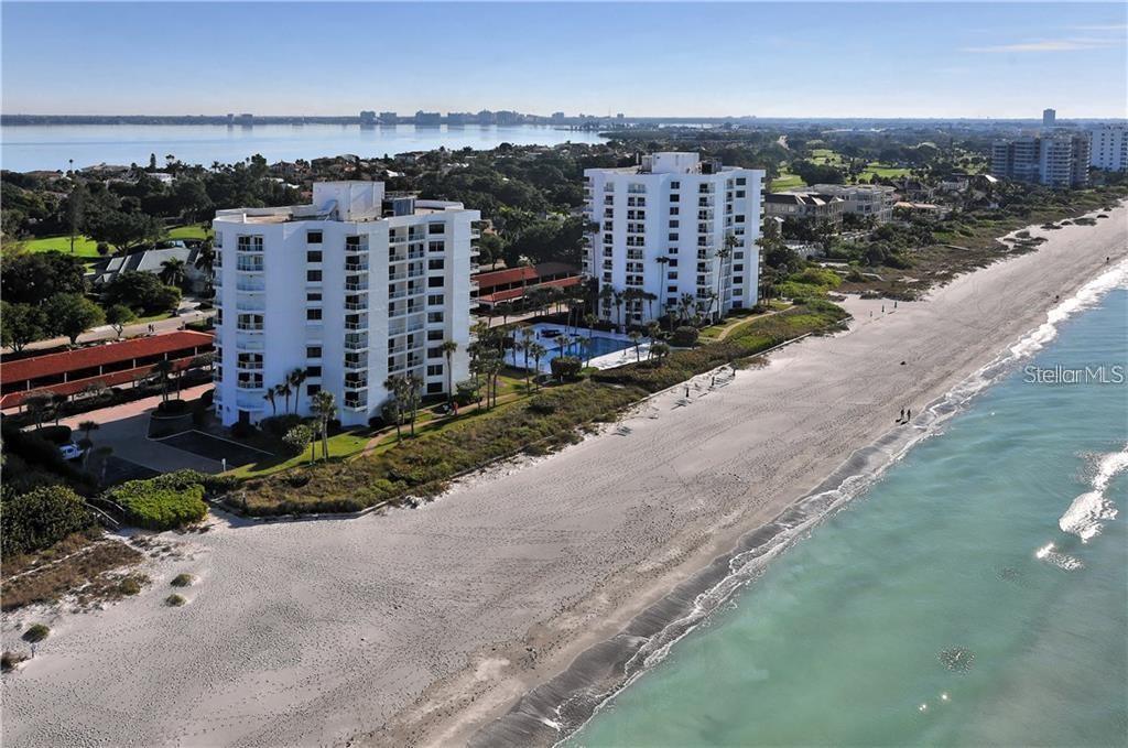An aerial view shows two white high-rise residential buildings situated directly on a sandy beach — 1050 LONGBOAT CLUB ROAD UNIT 502, LONGBOAT KEY, FL