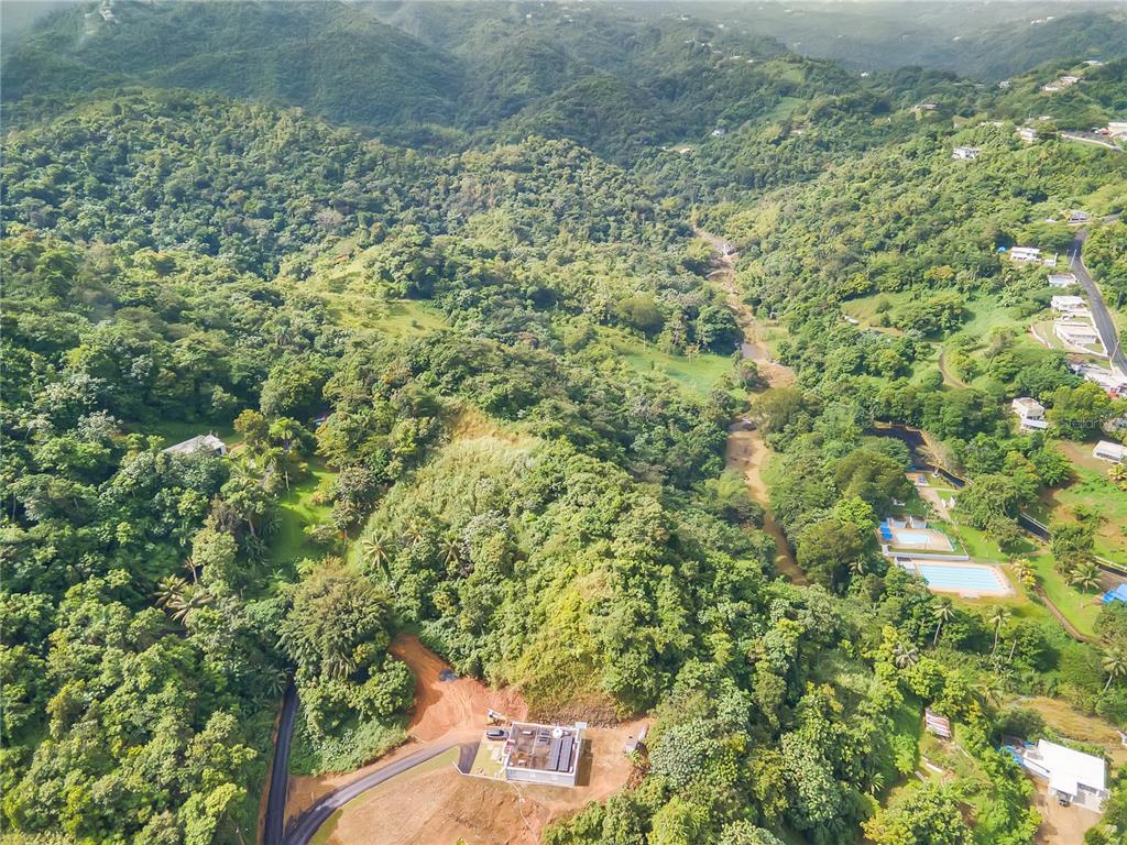 An aerial view shows a house under construction on a hillside surrounded by dense forest — BO. MULAS CARR. 174 KM. HM 19.6 INT. UNIT SOLAR 1, AGUAS BUENAS, PR