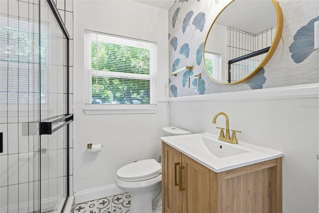 A bathroom features a wood vanity, gold faucet, round mirror, patterned wallpaper, and glass shower — 2855 VALENCIA WAY S, ST PETERSBURG, FL