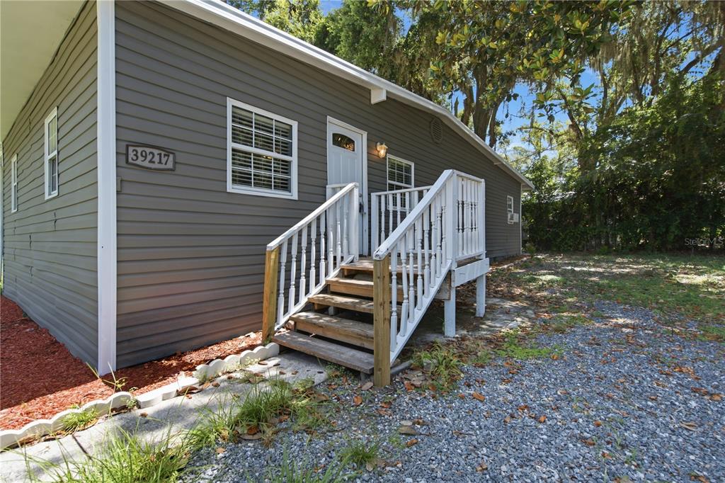 A gray-sided house exterior features a white front door with stairs and a wooden railing — 39217 FIBELKORN ROAD, ZEPHYRHILLS, FL