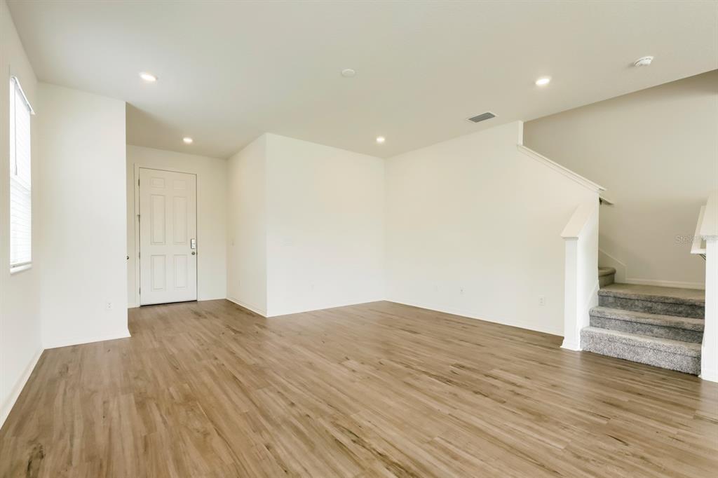 An empty living room features light wood-look flooring, white walls, a front door, and stairs — 37434 YUKON DRIVE, ZEPHYRHILLS, FL
