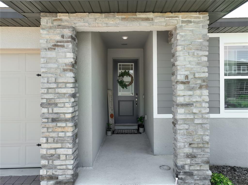 A stone-columned entryway leads to a gray front door with a wreath and welcome sign — 2327 BEAR PEAK ROAD, MINNEOLA, FL