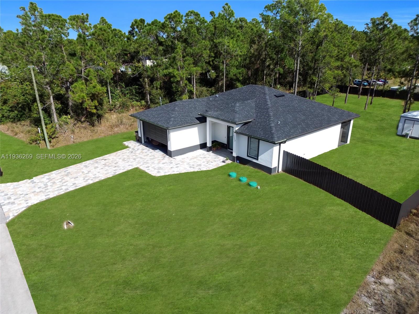 An aerial view shows a white house with a dark roof, paved driveway, and lawn — 1112 Clark Ave, Lehigh Acres FL 33972, LEHIGH ACRES, FL