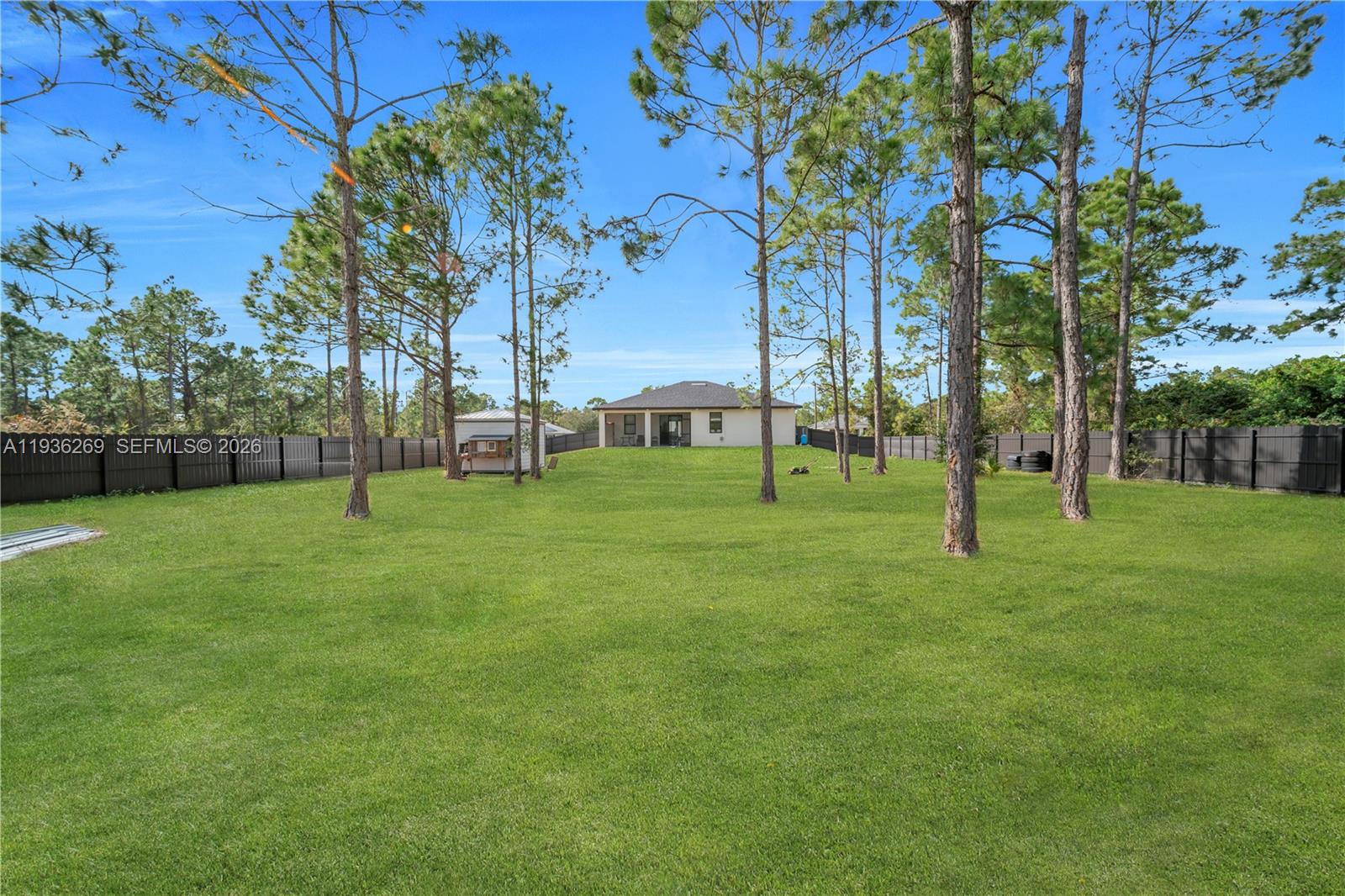 A large, fenced backyard features a grassy lawn, several tall pine trees, and a house — 1112 Clark Ave, Lehigh Acres FL 33972, LEHIGH ACRES, FL