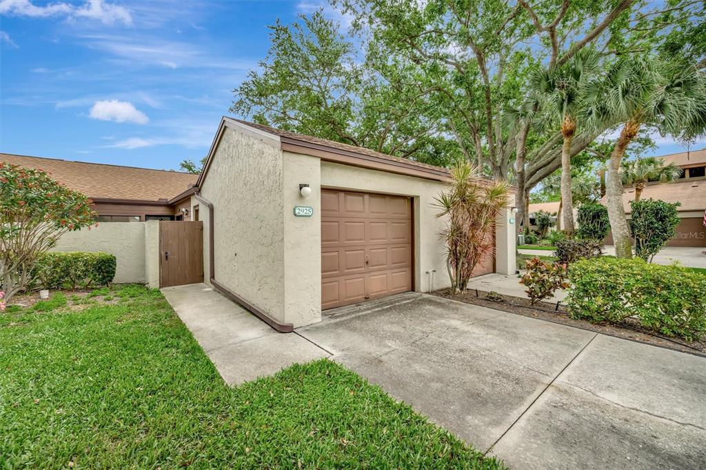 A tan stucco garage with a brown door and a concrete driveway under large trees — 2925 PARK LAKE DRIVE UNIT 12, BRADENTON, FL