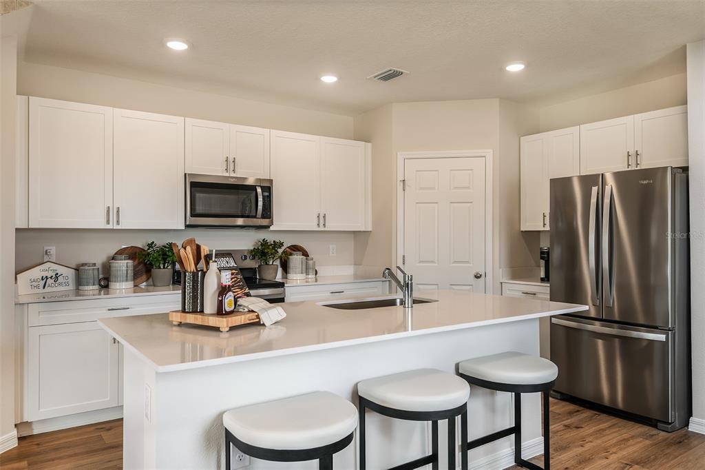 A modern kitchen features white cabinets, stainless steel appliances, a quartz island, and bar seating — 9864 PELICAN COAST DRIVE, DADE CITY, FL