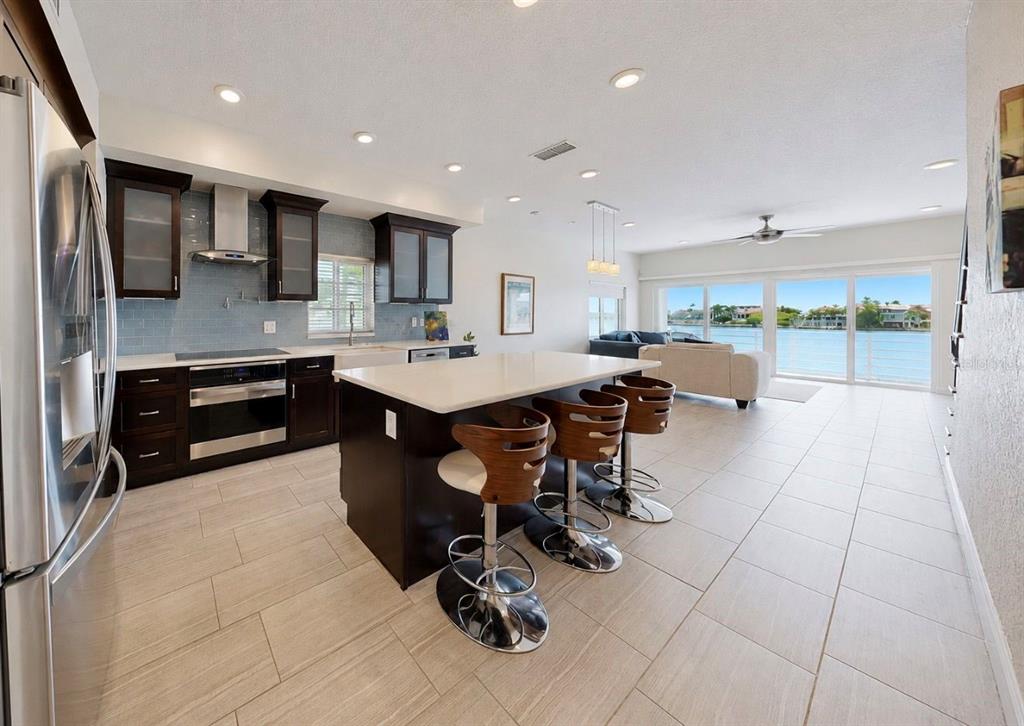 A modern kitchen with dark cabinets and a central island overlooks a waterfront living area — 4639 MIRABELLA COURT, ST PETE BEACH, FL