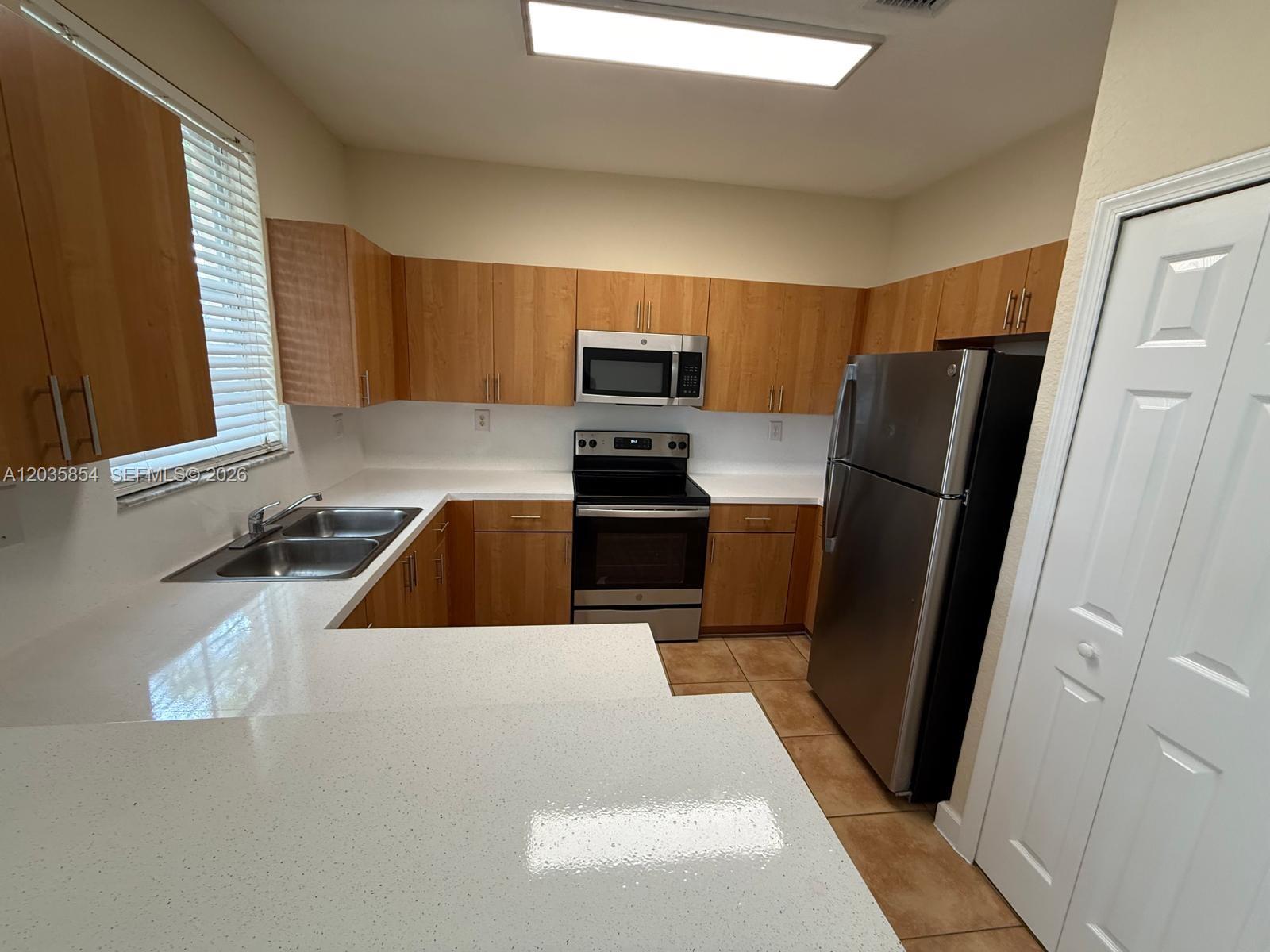 A kitchen features light wood cabinets, white countertops, stainless steel appliances, and tile flooring — 2462 SE 15th Ct, Homestead FL 33035, HOMESTEAD, FL