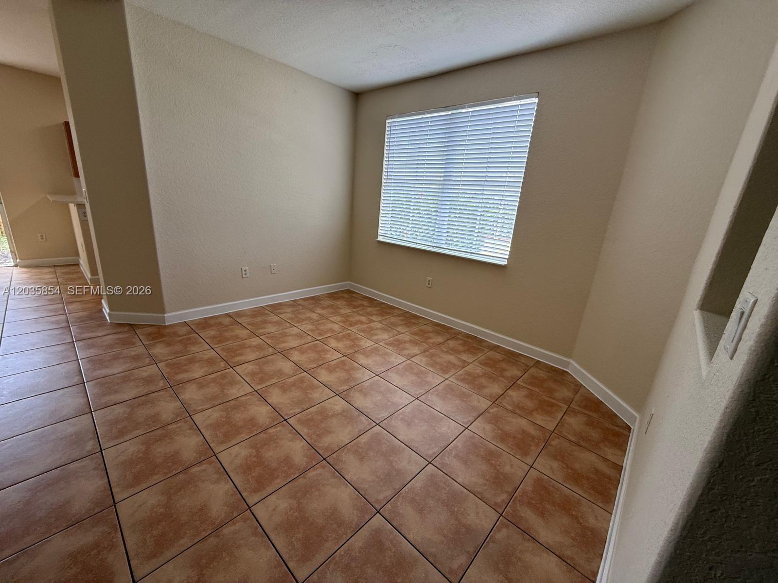 An empty room features tan walls, brown tile flooring, and a window with white blinds — 2462 SE 15th Ct, Homestead FL 33035, HOMESTEAD, FL