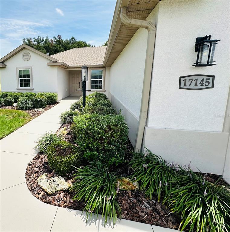 The exterior of a house features a tan stucco facade, a sidewalk, and landscaped bushes — 17145 SE 113TH AVENUE, SUMMERFIELD, FL