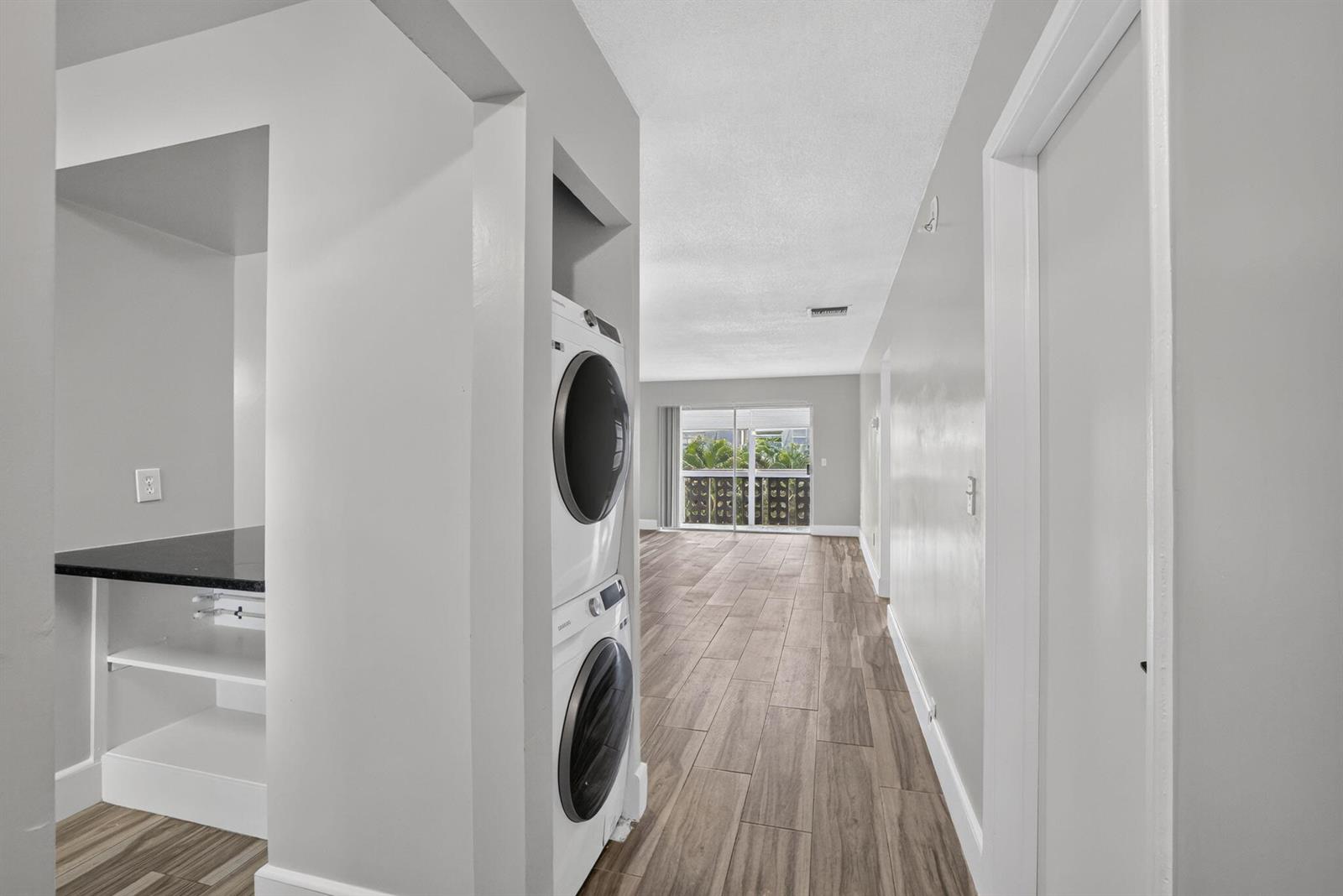 A hallway features wood-look tile flooring, a stacked washer and dryer, and a distant balcony — 801 Pine Drive # 12, Pompano Beach FL 33060, POMPANO BEACH, FL