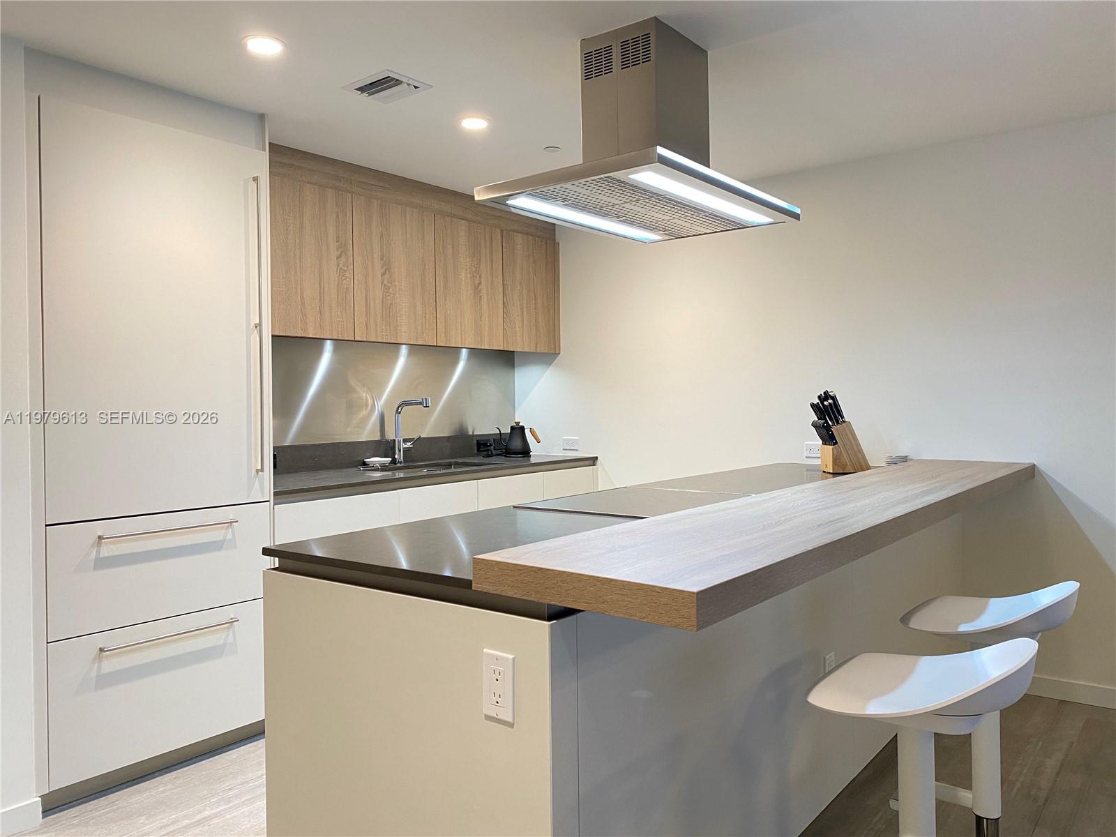 A modern kitchen features white cabinetry, wood-toned upper cabinets, a dark island, and a range hood — 1000 Brickell Plaza # 3807, Miami FL 33131, MIAMI, FL