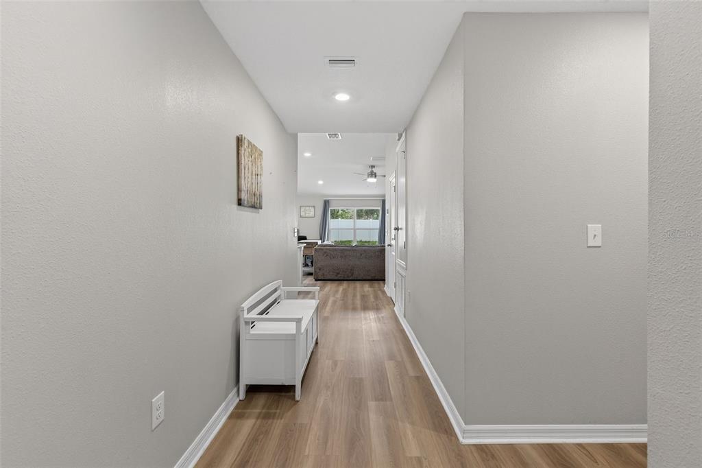 A hallway with light wood-look flooring leads to a living area with a gray sofa — 9972 MESIC WOODS AVENUE, SEFFNER, FL