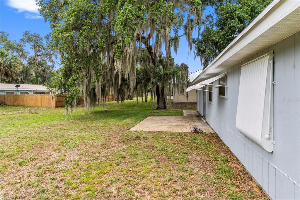 A side view of a house with light blue siding, a concrete patio, and large trees — 326 BELFAST TERRACE, SEBASTIAN, FL