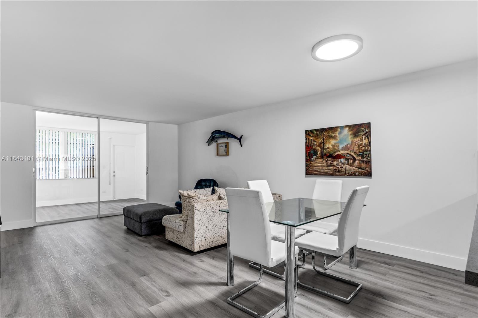 A dining area with a glass table and white chairs sits on gray wood flooring — 445 NE 195th St # 125, Miami FL 33179, MIAMI, FL