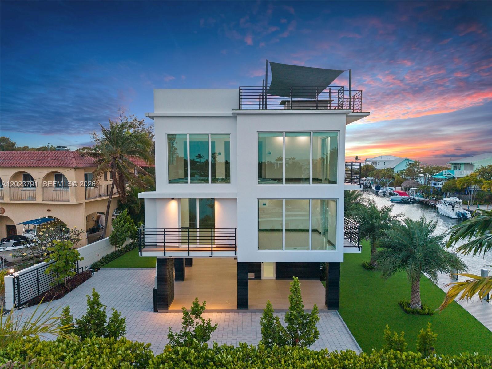 A modern three-story white house with large windows sits on stilts beside a canal — 265 Pueblo St, Tavernier FL 33070, TAVERNIER, FL