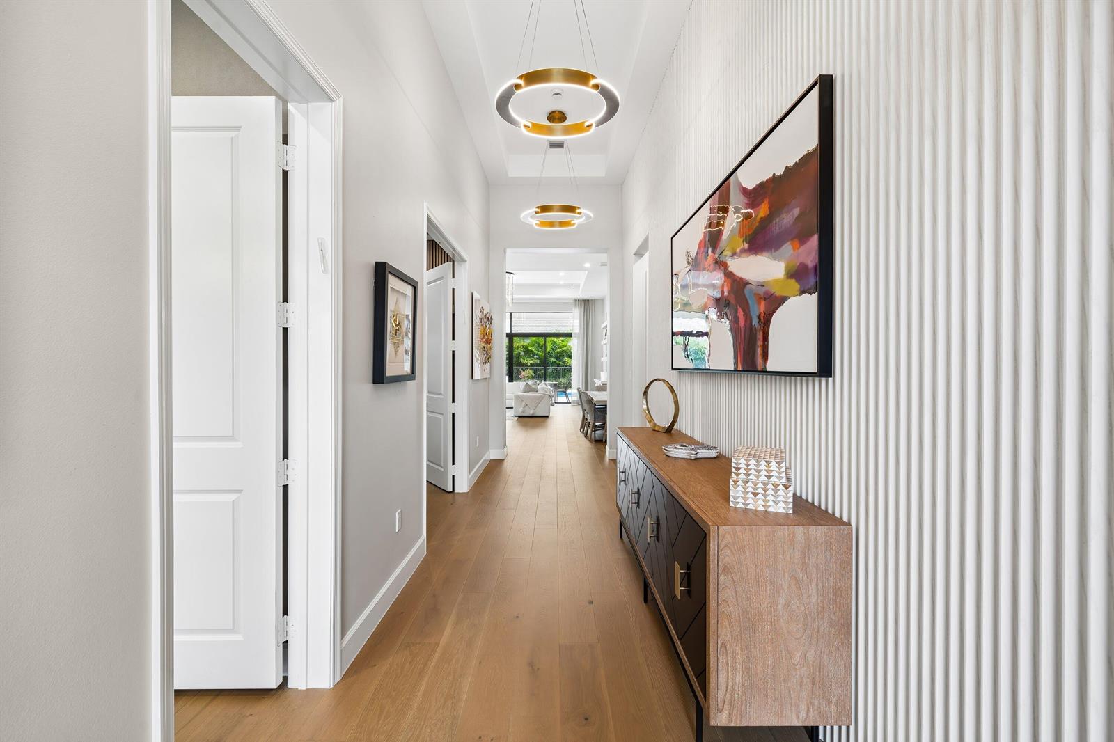 A hallway features wood flooring, two gold ring chandeliers, and a textured white accent wall — 9812 Spruce Woods Drive, Boynton Beach FL 33473, BOYNTON BEACH, FL