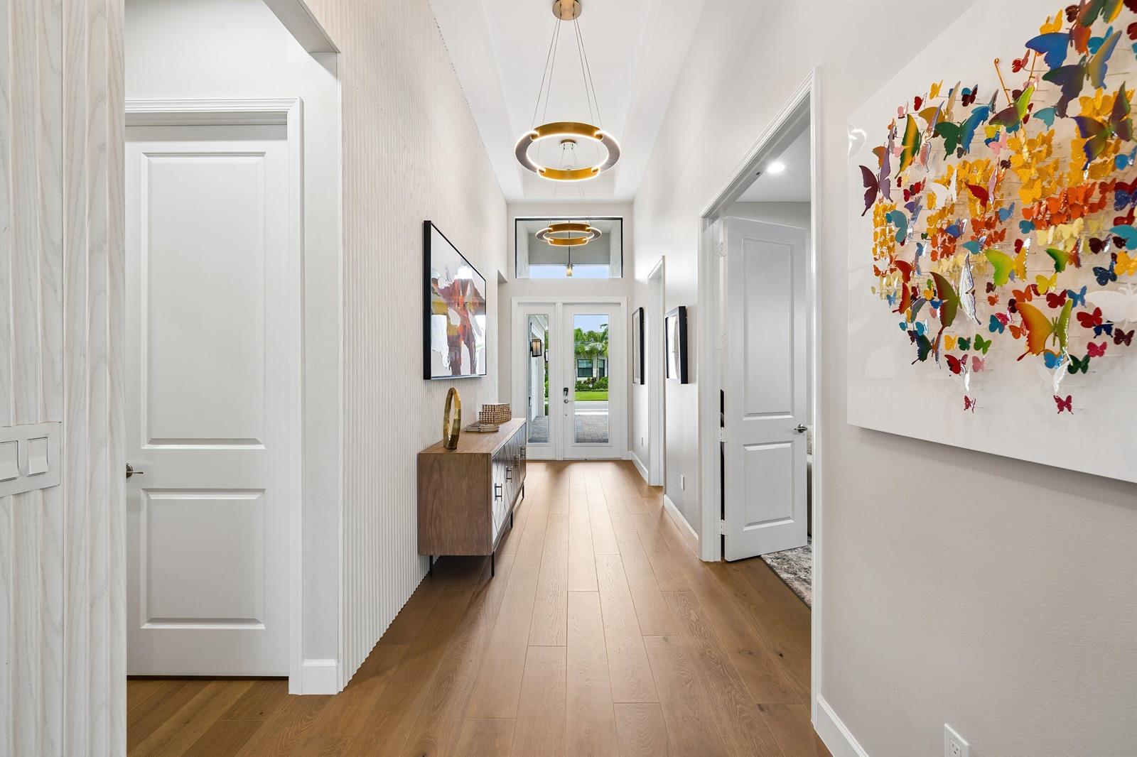 A bright hallway features light wood floors, two circular chandeliers, and a colorful butterfly wall art — 9812 Spruce Woods Drive, Boynton Beach FL 33473, BOYNTON BEACH, FL