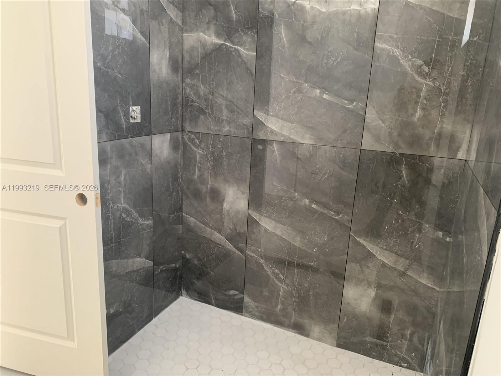 A shower stall features large-format gray marble-look wall tiles and a white hexagonal floor — 1016 Cortez Ave, Lehigh Acres FL 33974, LEHIGH ACRES, FL