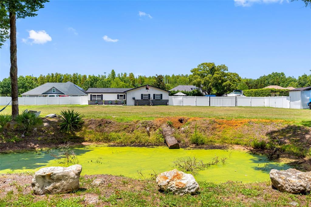 A backyard features a green pond in the foreground and a white-fenced house behind — 5515 LAKE LE CLARE ROAD, LUTZ, FL