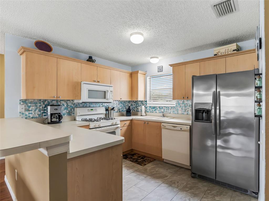 A kitchen features light wood cabinets, stainless steel appliances, and a blue mosaic tile backsplash — 897 LIVINGSTON LOOP, THE VILLAGES, FL