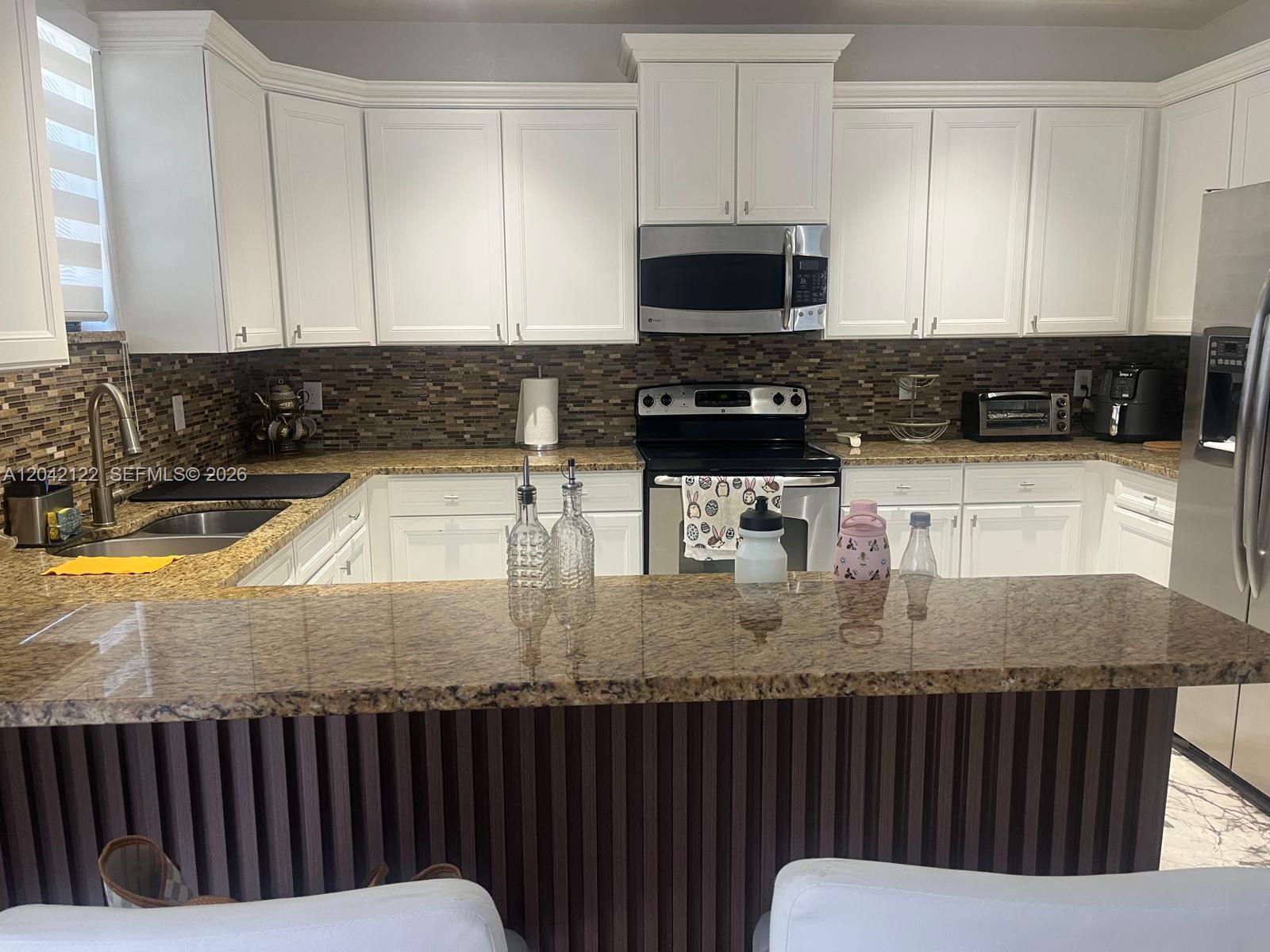 A kitchen features white cabinets, granite countertops, a brown tiled backsplash, and a breakfast bar — 8985 NW 98th Ave, Doral FL 33178, DORAL, FL