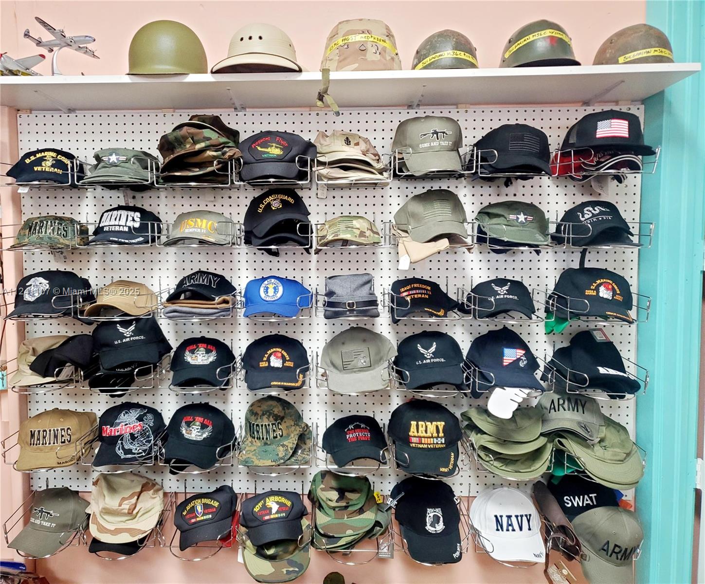 A wall-mounted display rack holds numerous military-themed hats and helmets in a room — 755 NW Federal Hwy, Stuart FL 34997, STUART, FL