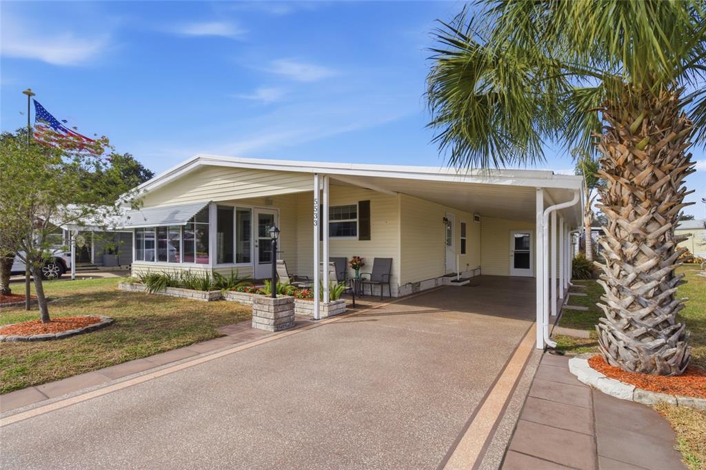 A light yellow manufactured home features a covered carport, a front porch, and a palm tree — 5533 REGAL WAY, ZEPHYRHILLS, FL