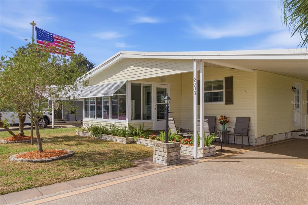 A light yellow manufactured home with a covered carport, front porch, and American flag — 5533 REGAL WAY, ZEPHYRHILLS, FL