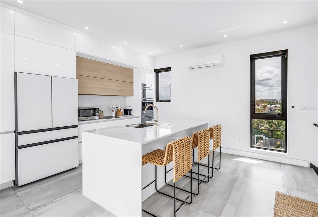 A modern kitchen features white cabinetry, a marble-topped island with woven stools, and large windows — 30 80TH AVENUE, TREASURE ISLAND, FL