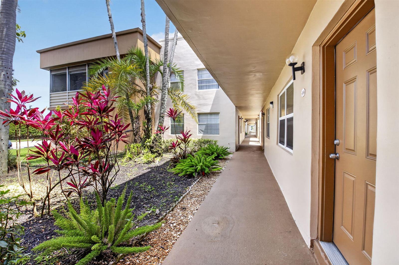 An outdoor walkway leads past a tan apartment building with tropical landscaping and a brown door — 14 Flanders # A, Delray Beach FL 33484, DELRAY BEACH, FL