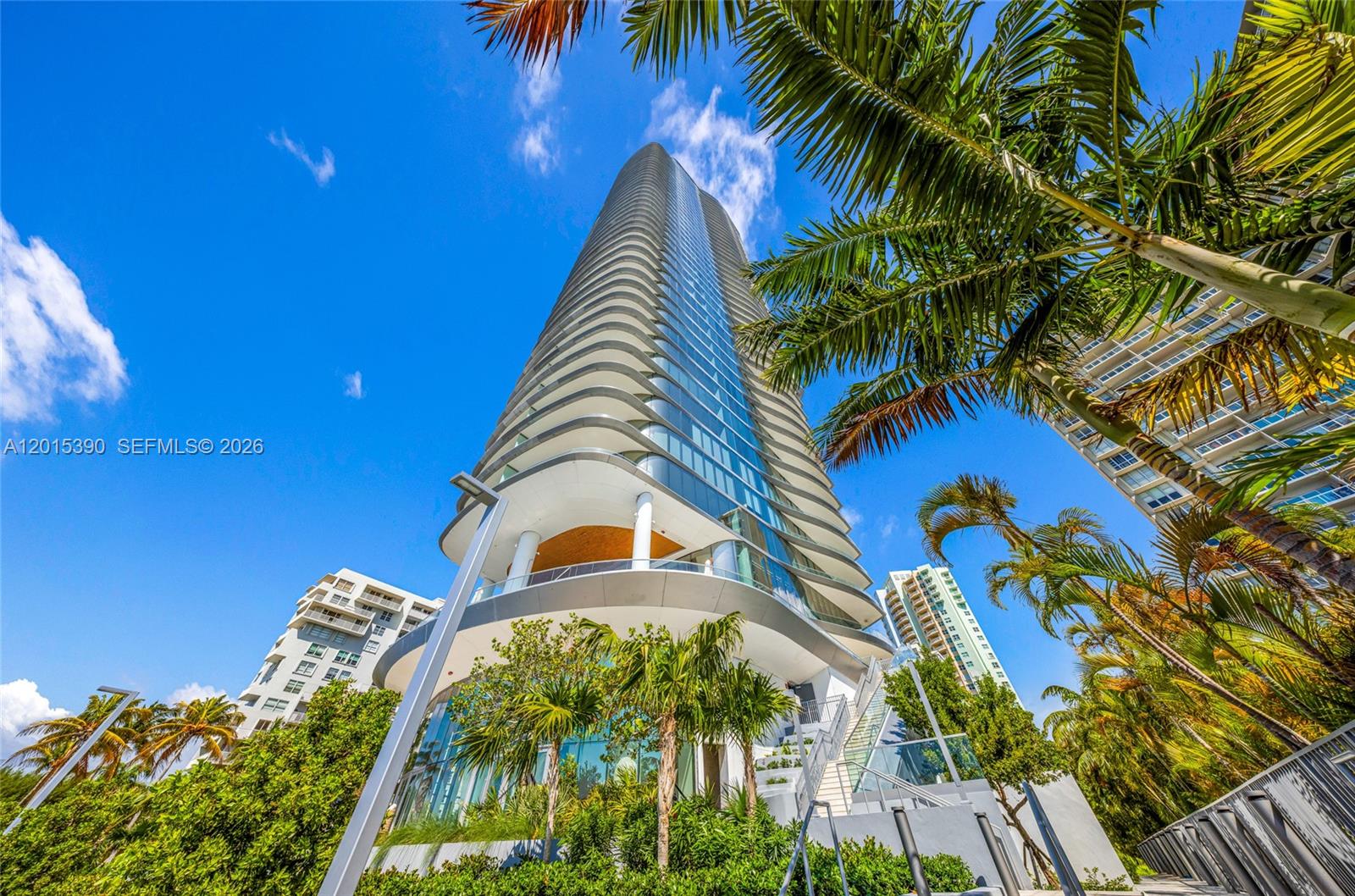 A low-angle view of a modern, curved glass high-rise building surrounded by palm trees — 175 SE 25 Road # 1203, Miami FL 33129, MIAMI, FL